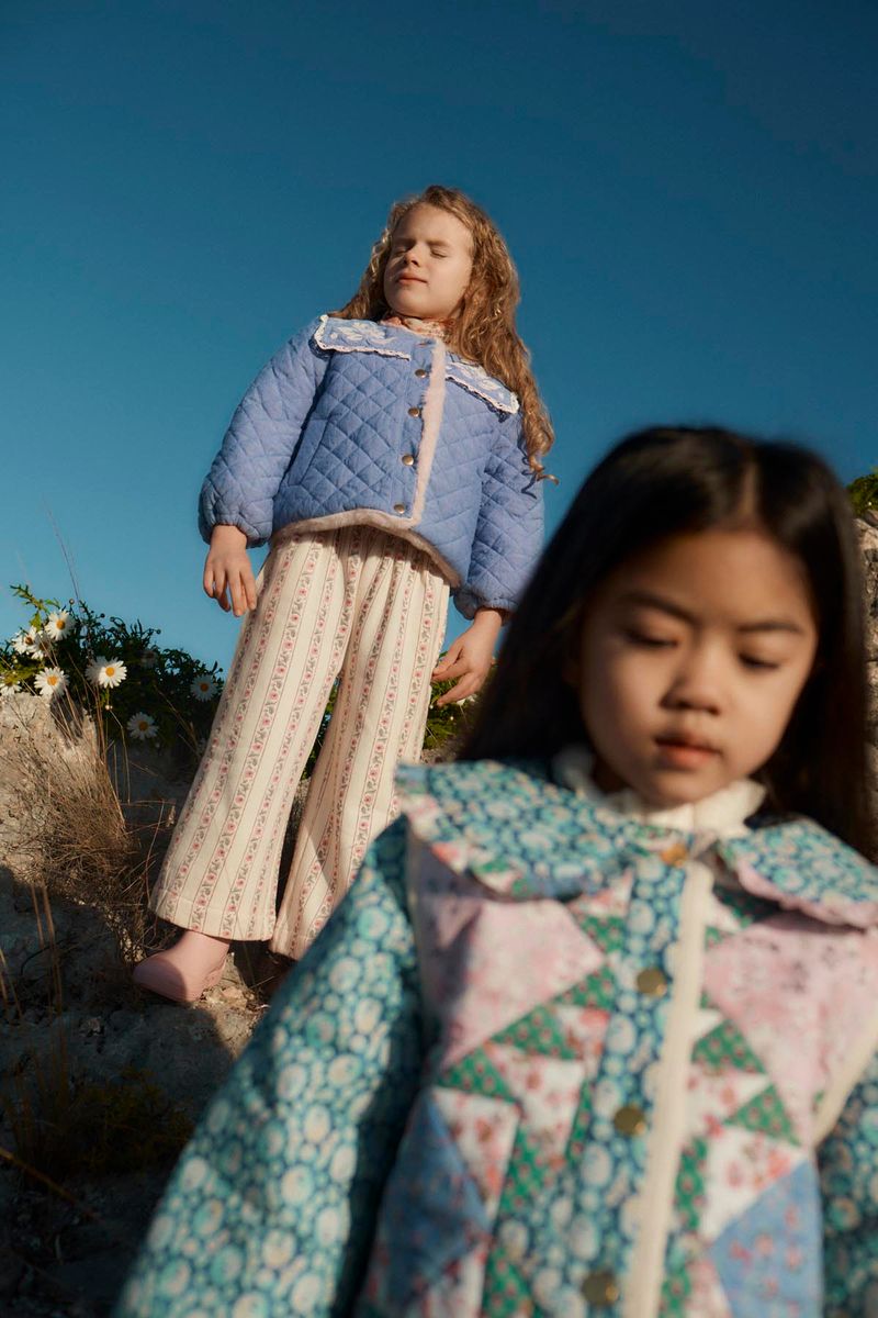 Two young girls stand outdoors on a rocky slope, both wearing patterned jackets and the Louise Misha Girls’ Ysalia Sweatpant with an elasticated waist, under a clear blue sky.