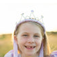 A smiling young girl wearing a purple princess dress and a Great Pretenders Ribbon Tiara Pink stands outdoors in the sunlight.