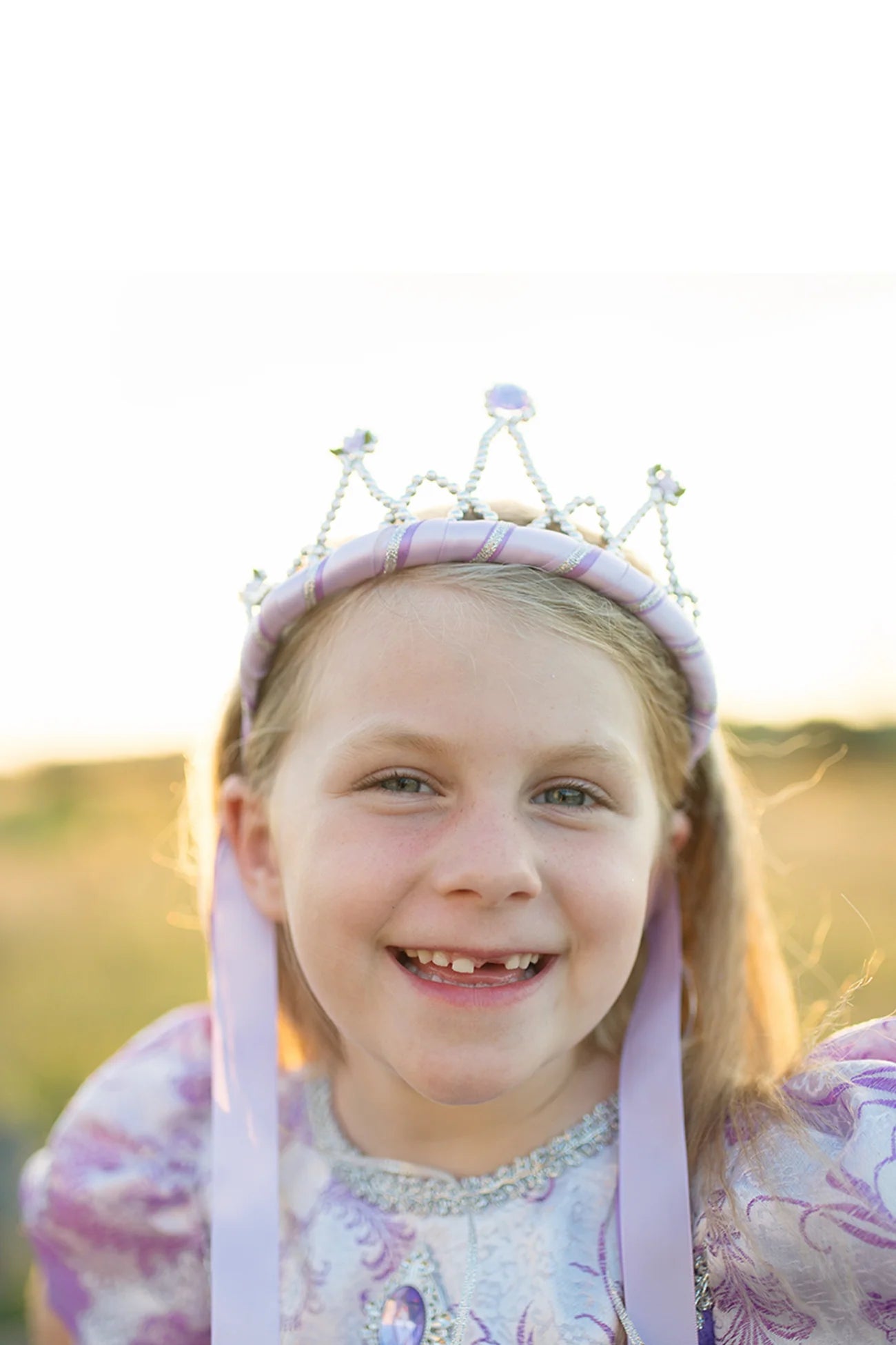 A smiling young girl wearing a purple princess dress and a Great Pretenders Ribbon Tiara Pink stands outdoors in the sunlight.