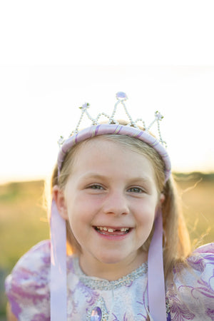 A smiling young girl wearing a purple princess dress and a Great Pretenders Ribbon Tiara Pink stands outdoors in the sunlight.