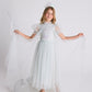 A young girl beams as she poses against a plain background, wearing the Tutu du Monde Girls’ Snow Queen Tutu Dress paired with a sparkling tiara and wand, elegantly spreading the sheer tulle wings of her magical ice queen costume.