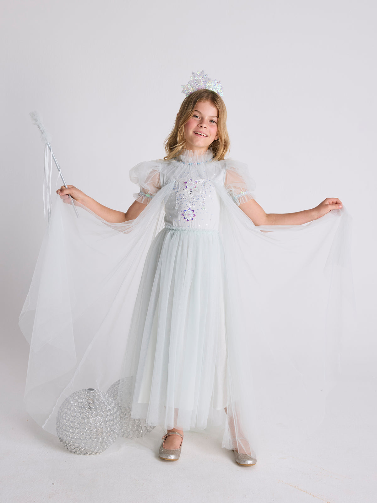 A young girl beams as she poses against a plain background, wearing the Tutu du Monde Girls’ Snow Queen Tutu Dress paired with a sparkling tiara and wand, elegantly spreading the sheer tulle wings of her magical ice queen costume.