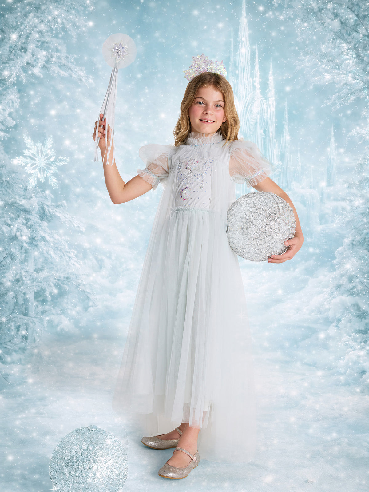 A girl in a Snow Queen Tutu Dress holds the sparkling Tutu du Monde Snow Queen Wand and a decorative ball, standing among glistening icicles in a magical snowy, icy fantasy setting.