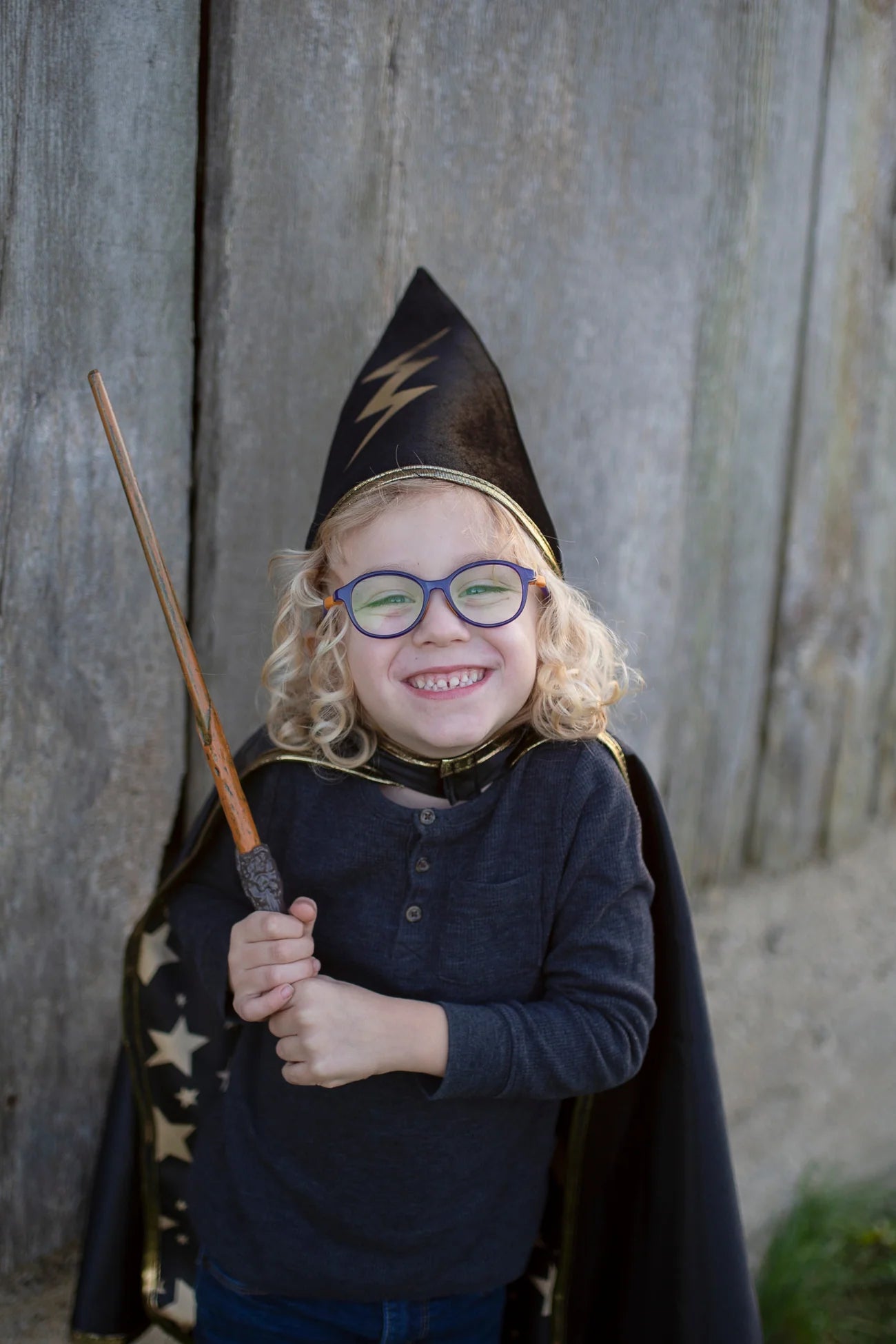 A smiling child in glasses wears the Great Pretenders Reversible Wizard Cape & Hat and holds a wand while standing in front of a wooden wall.