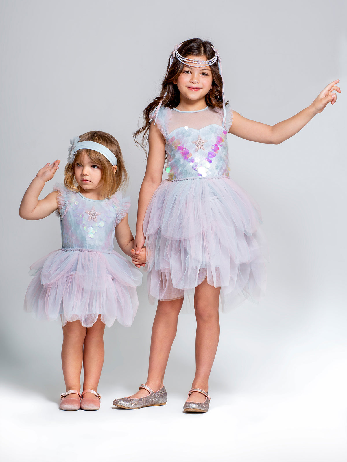 Two young girls wearing Tutu du Monde Baby Girls’ Bebe Mermaid Magic Tutu Dresses stand against a plain background, each with a matching headband, holding hands and raising one arm.