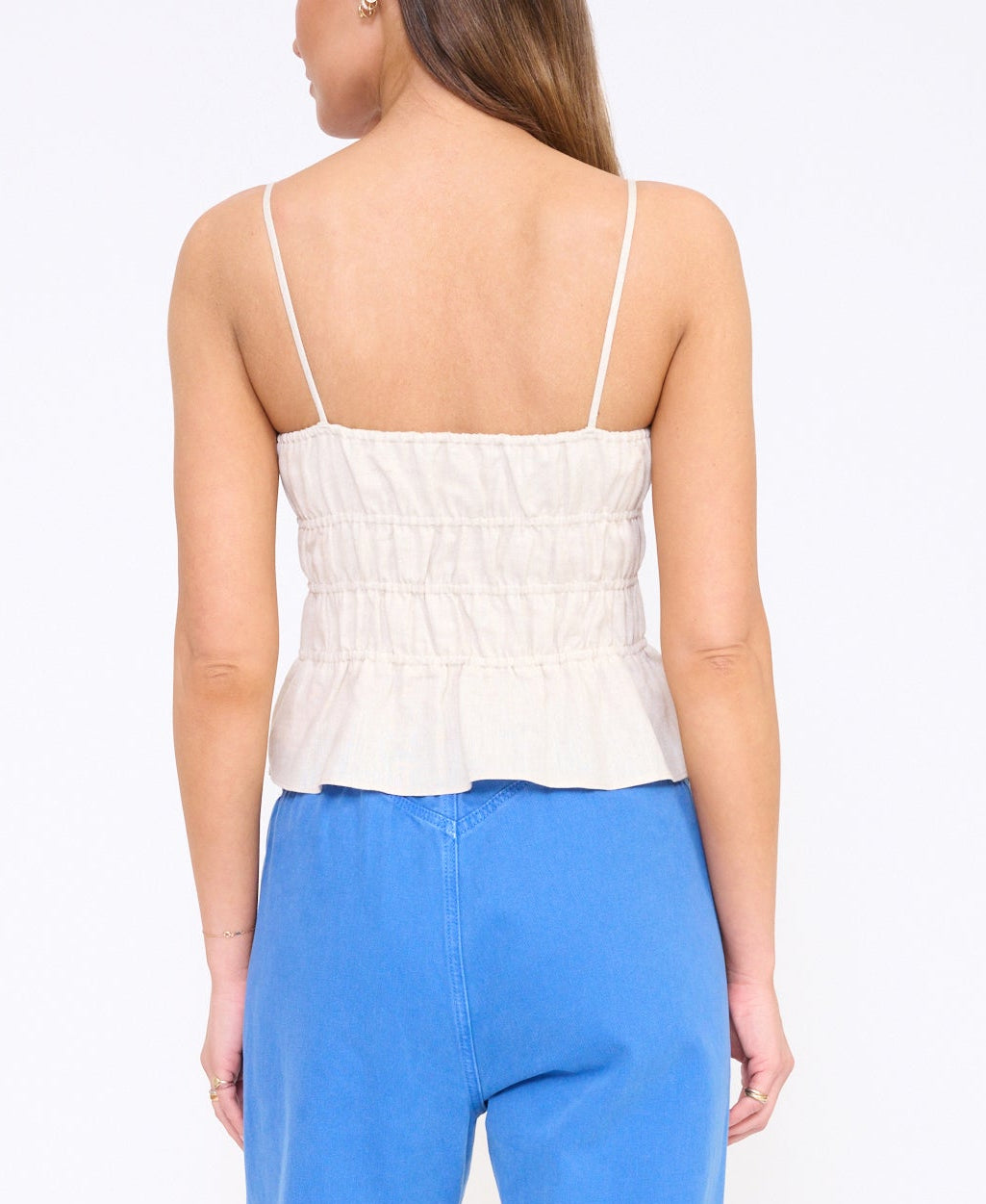 A woman is shown from the back against a plain light background, wearing the Staud Ayla Linen Tank—a sleeveless cami with thin straps—and blue high-waisted pants with a cropped silhouette.
