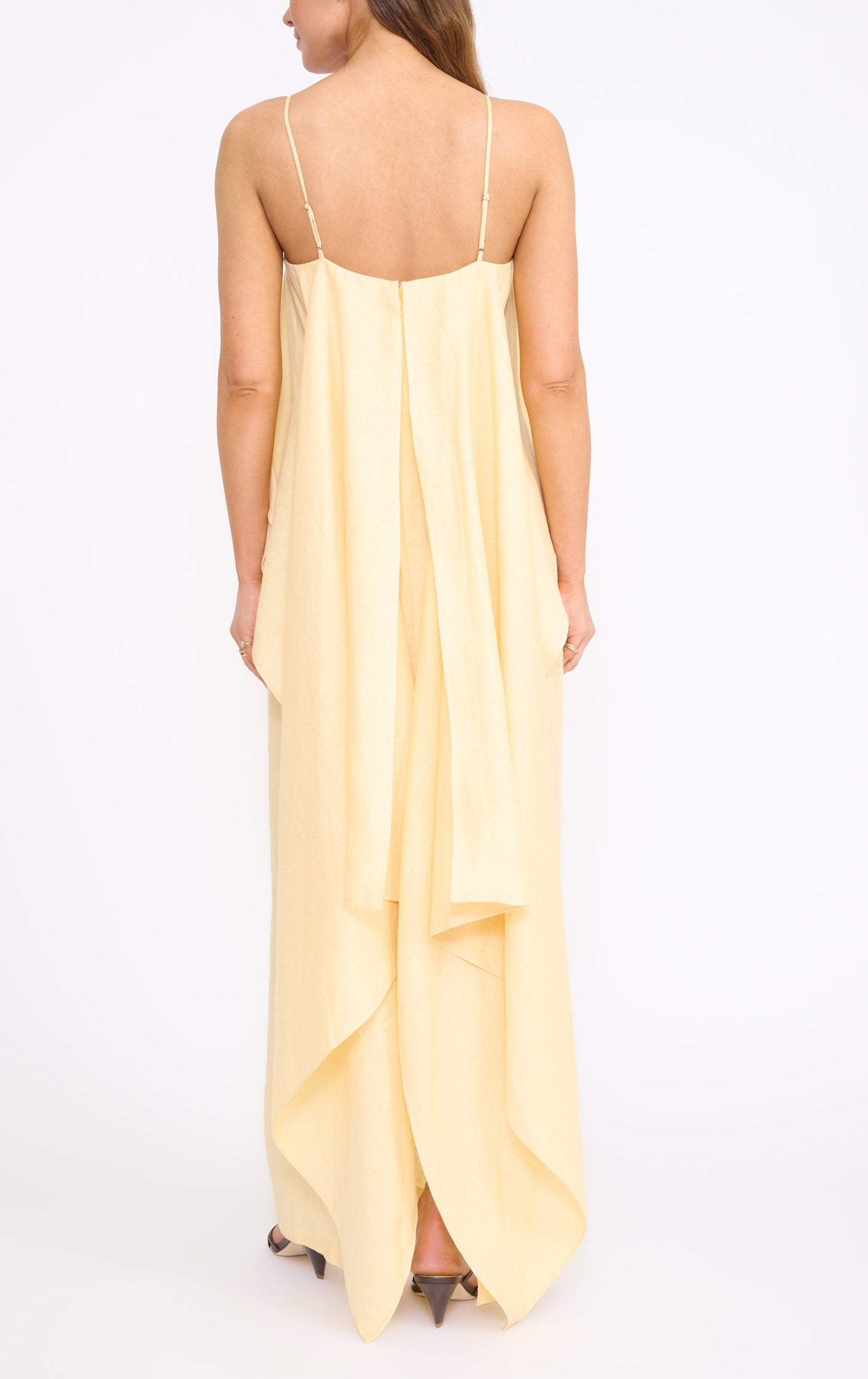 A woman faces away in the Staud Valencia Linen Dress, a light yellow style with adjustable straps and a draped, layered back that enhances its elegant column silhouette.