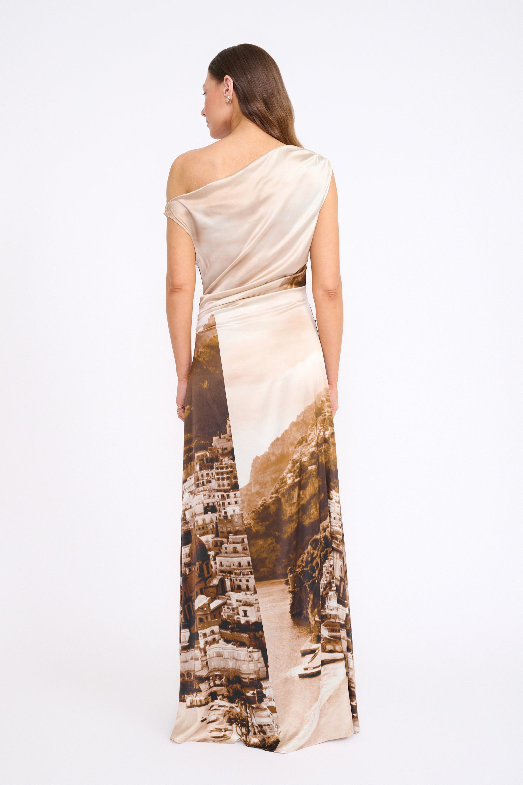 A woman stands facing away, wearing the Staud Phare Silk Maxi Dress featuring a one-shoulder neckline and a scenic city and coastline print on the skirt.