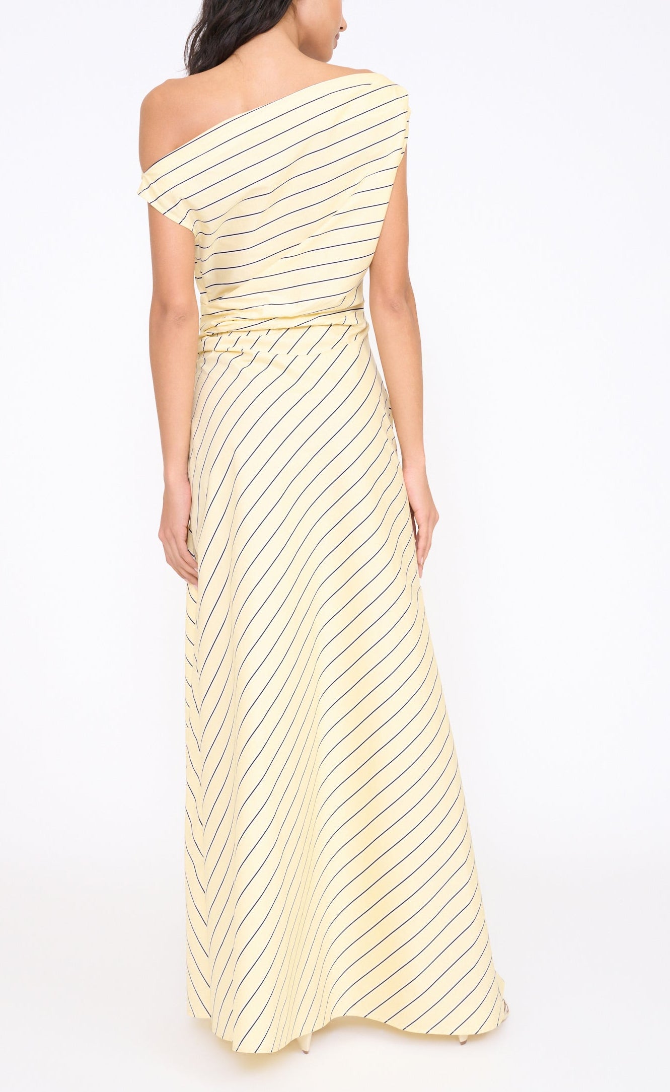 A woman is seen from behind in the Staud Phare Maxi Dress, a floor-length yellow piece with black diagonal stripes and a one-shoulder design, the printed fabric flowing elegantly against a plain white background.