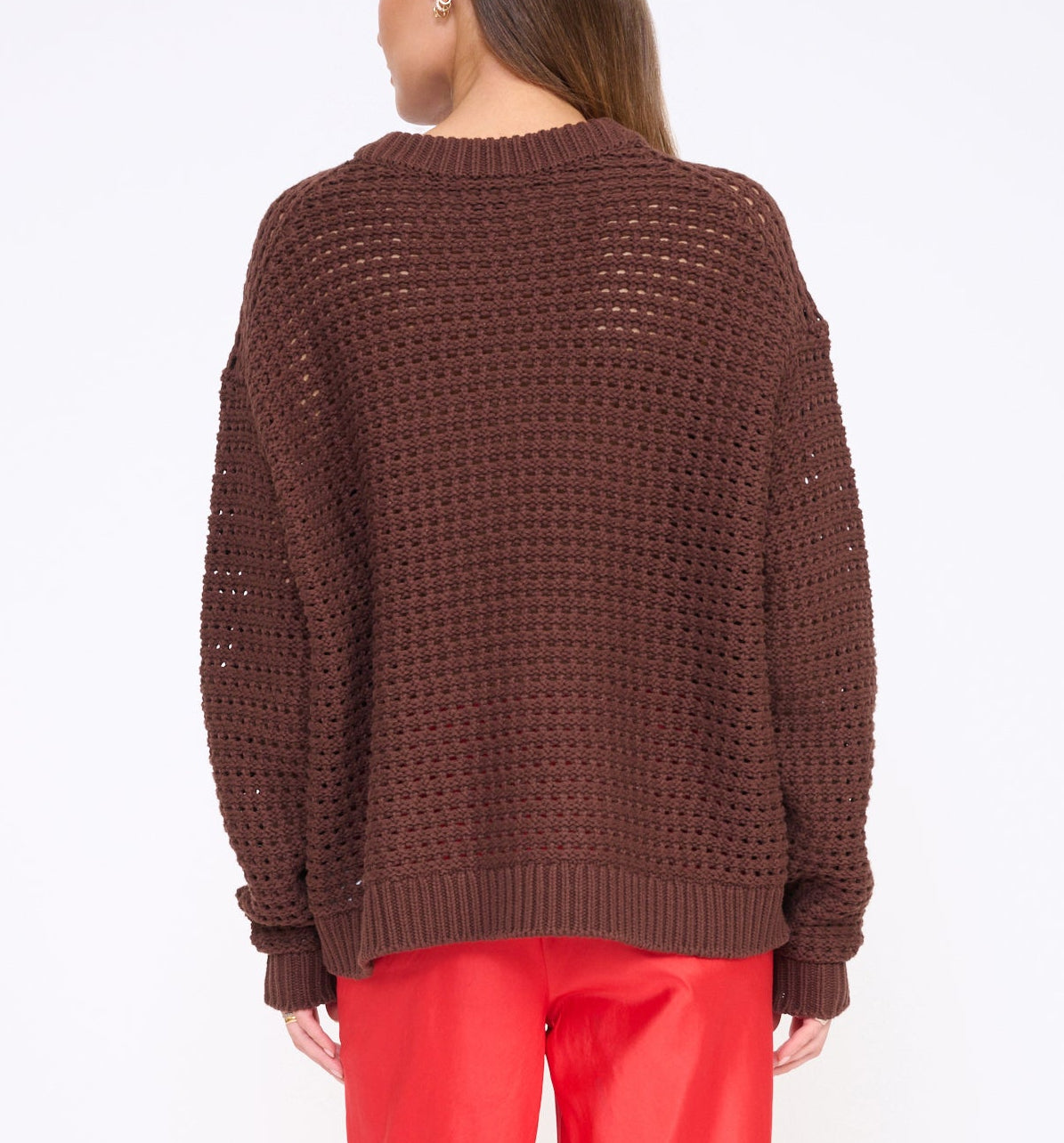 A person stands with their back to the camera against a plain white background, wearing Staud's Giada Seahorse Sweater in a loose, boxy fit with drop shoulders, paired with red pants.