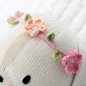 A close-up of Cuddle + Kind’s Charlotte the Dog - Regular 20" wearing a pink and peach wildflowers headband with green leaves, set against a white textured background.