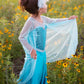 A young child in the Great Pretenders Ice Queen Dress With Cape, featuring a sequin sweetheart bodice, stands among yellow wildflowers and gazes to the side while holding out their shimmering blue skirt.