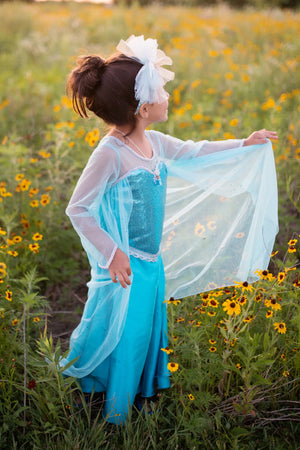 A young child in the Great Pretenders Ice Queen Dress With Cape, featuring a sequin sweetheart bodice, stands among yellow wildflowers and gazes to the side while holding out their shimmering blue skirt.