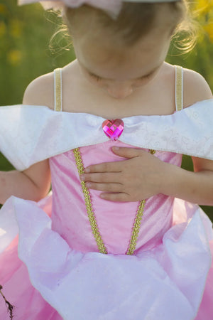 A child wearing the Great Pretenders Deluxe Sleeping Cutie Gown, a pink princess dress with gold trim and a heart-shaped jewel, holds a hand to their chest while looking down.