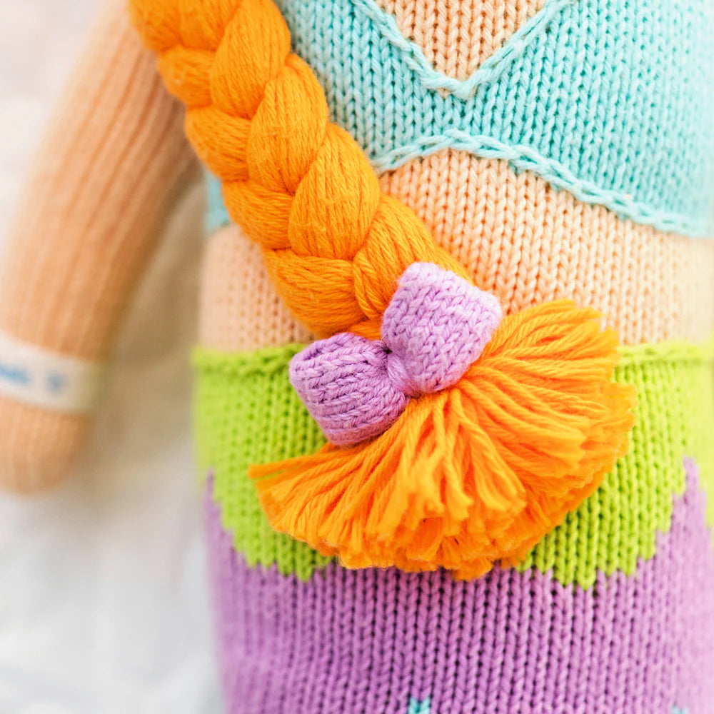 Close-up of Isla the Mermaid, a 13" knitted doll by Cuddle + Kind, with orange braided yarn hair tied with a purple bow and dressed in a blue-green top and purple skirt—ready to spread kindness in her magical world.