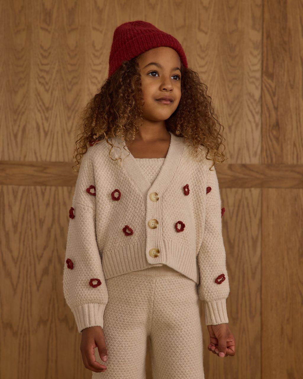 A young child with curly hair wears the Rylee & Cru Baby Girls' and Girls' Boxy Crop Cardigan, standing in front of a wooden wall.