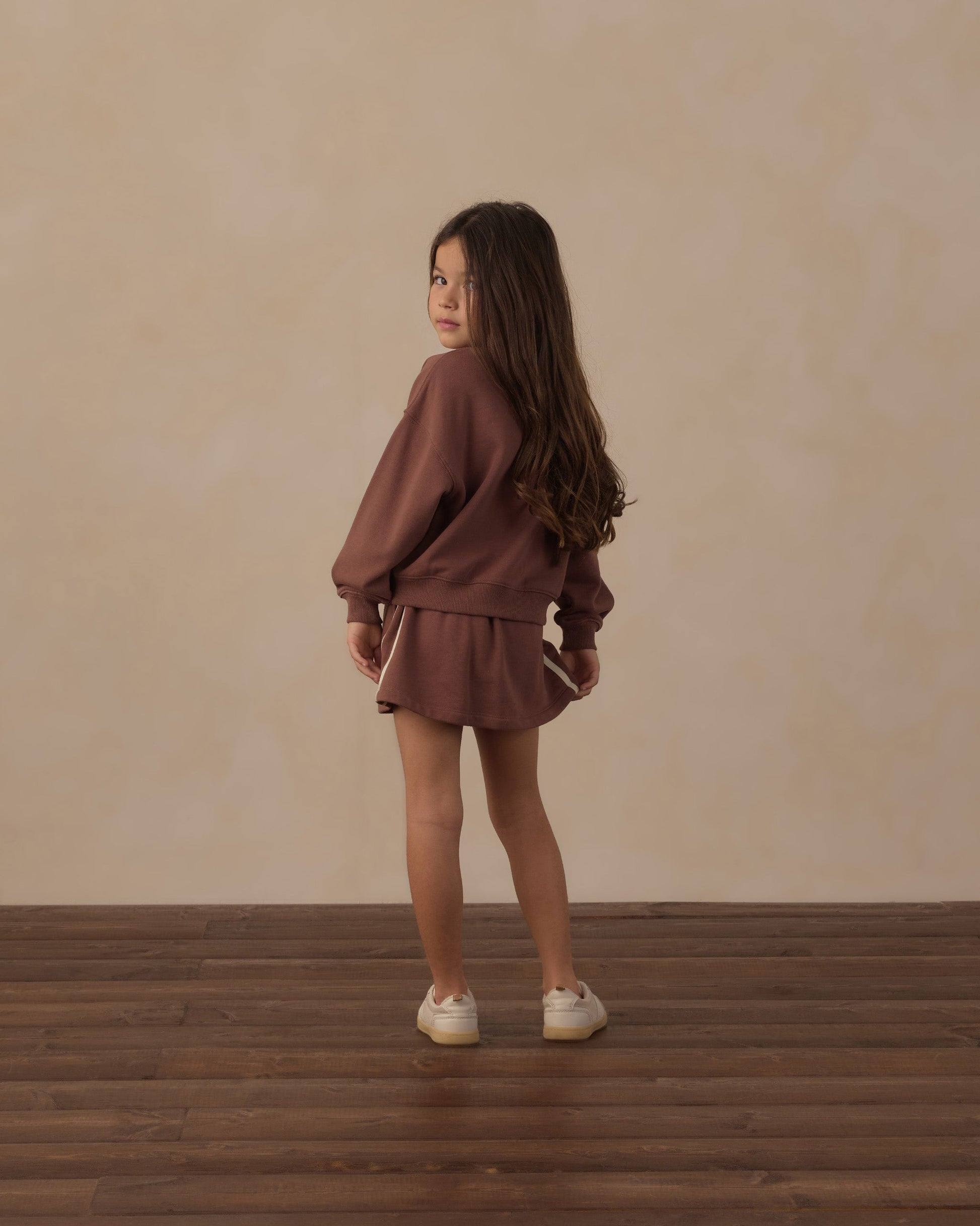 A young girl with long brown hair stands on a wooden floor, wearing the Rylee & Cru Girls’ Sports Skirt with an elastic waistband and a coordinating brown long-sleeve shirt, looking over her shoulder toward the camera for an effortlessly athletic look.