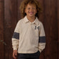 A young child with curly hair smiles while wearing the Rylee & Cru Baby Boys’ and Boys’ Rugby Polo—a cream long-sleeve shirt with a dark rugby stripe, classic collar, and number 14—standing in front of a wooden wall.
