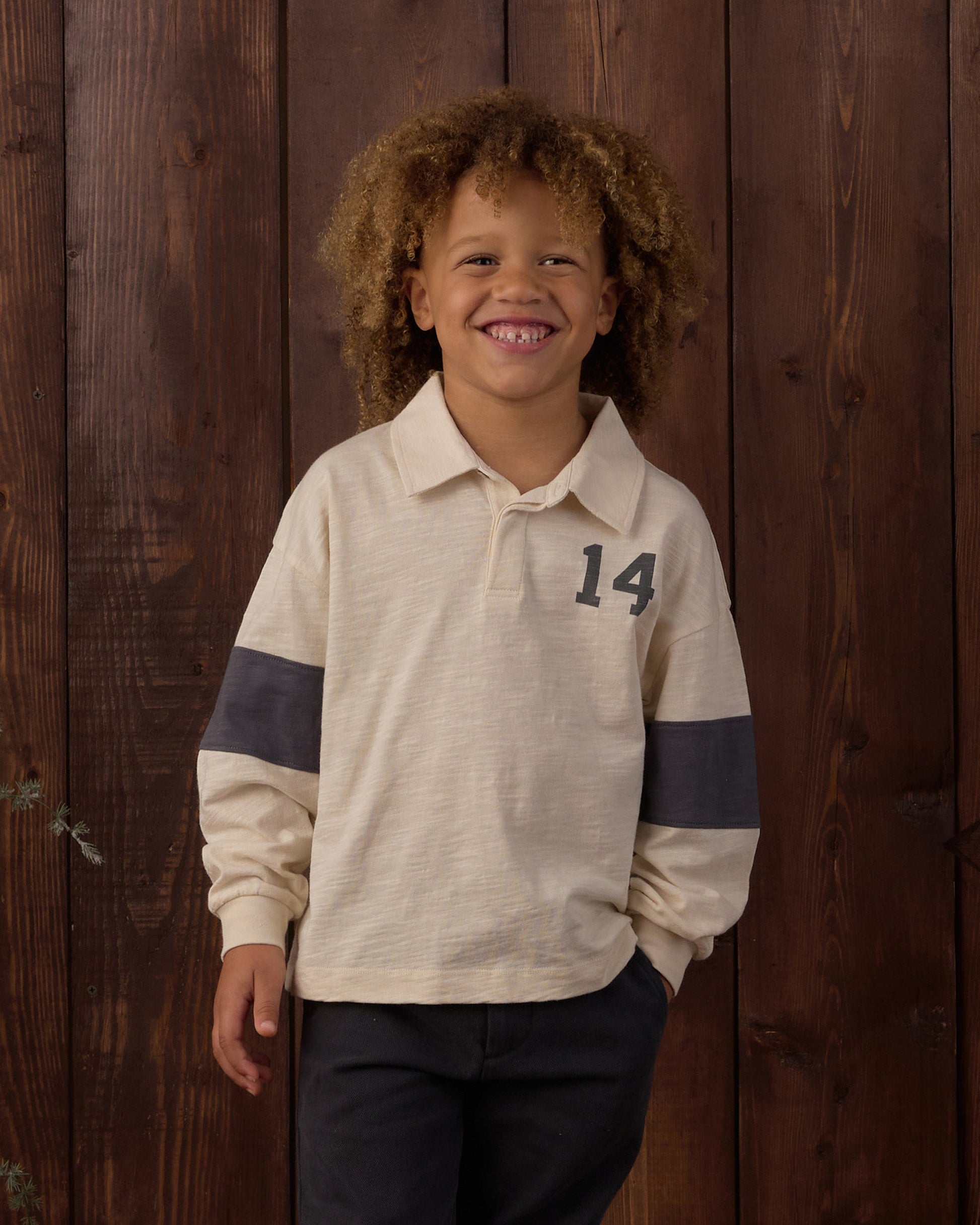 A young child with curly hair smiles while wearing the Rylee & Cru Baby Boys’ and Boys’ Rugby Polo—a cream long-sleeve shirt with a dark rugby stripe, classic collar, and number 14—standing in front of a wooden wall.