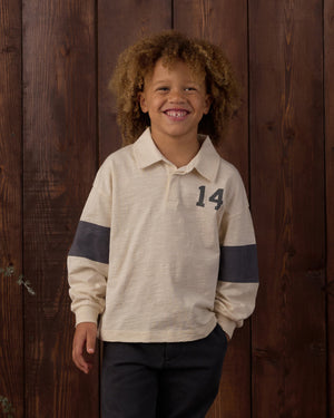 A young child with curly hair smiles while wearing the Rylee & Cru Baby Boys’ and Boys’ Rugby Polo—a cream long-sleeve shirt with a dark rugby stripe, classic collar, and number 14—standing in front of a wooden wall.