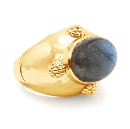 The Cleopatra Oval Ring by Juliska Capucine De Wulf features a wide gold band, a large oval blue labradorite stone, and decorative gold bead accents.