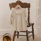 The Quincy Mae Baby Girls’ and Girls’ Ruffle Collar Jersey Dress, featuring a charming floral pattern perfect for everyday wear, hangs on a wooden hanger displayed over a wooden chair; nearby, a small basket holding three pears sits on the floor.