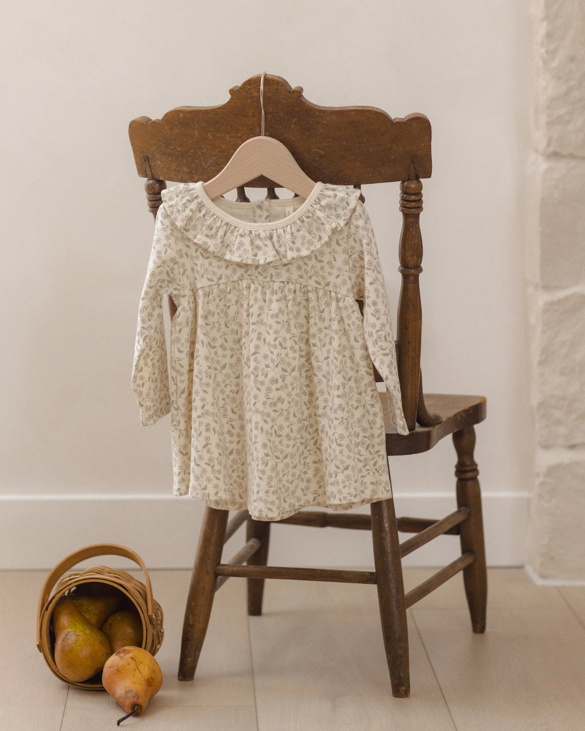 The Quincy Mae Baby Girls’ and Girls’ Ruffle Collar Jersey Dress, featuring a charming floral pattern perfect for everyday wear, hangs on a wooden hanger displayed over a wooden chair; nearby, a small basket holding three pears sits on the floor.