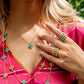 A woman in a pink top showcases gold jewelry with turquoise accents, featuring a Juliska Capucine De Wulf Berry Gem Ring, a necklace, and a long chain.