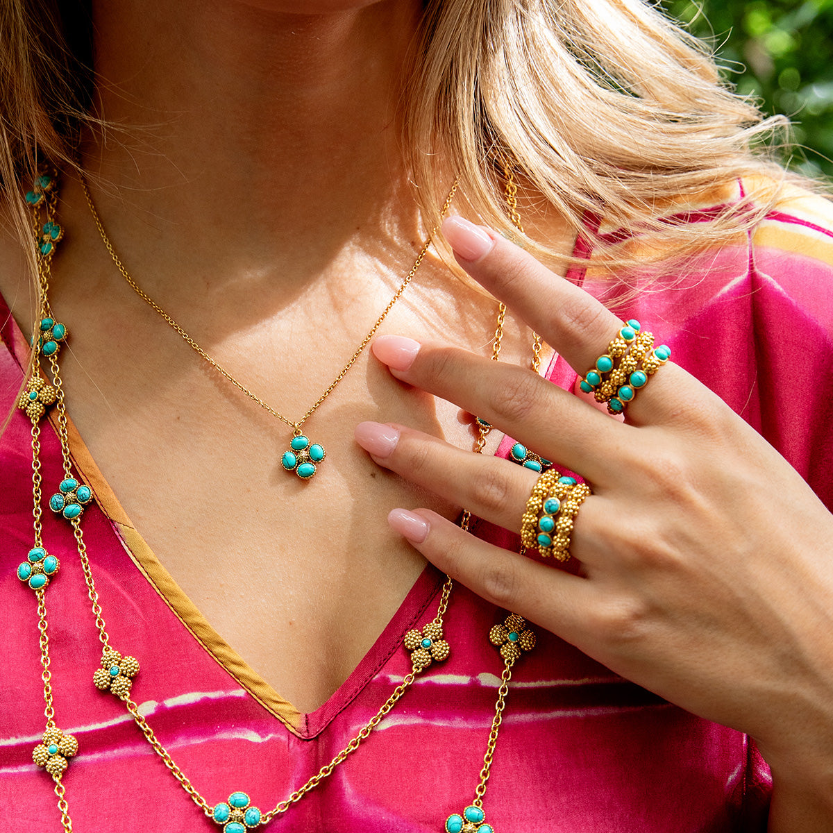 A woman in a pink top showcases gold jewelry with turquoise accents, featuring a Juliska Capucine De Wulf Berry Gem Ring, a necklace, and a long chain.