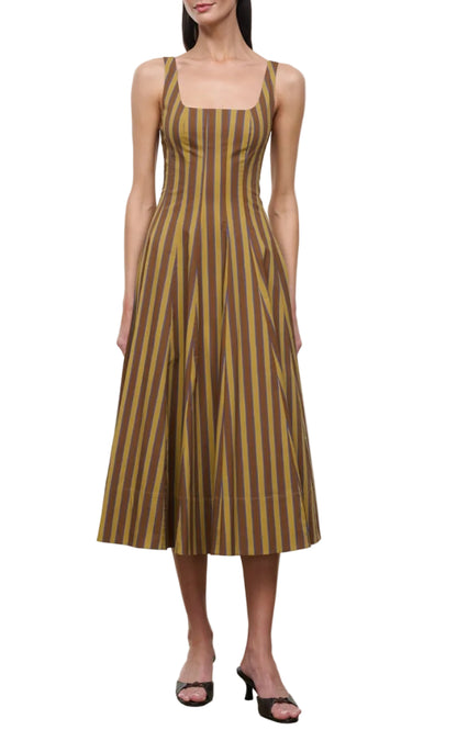 Wearing the Staud Wells Midi Cotton Poplin Dress, a sleeveless olive and brown striped midi dress with a square neckline, paired with block-heeled sandals, a woman stands against a white background, capturing the signature STAUD silhouette.
