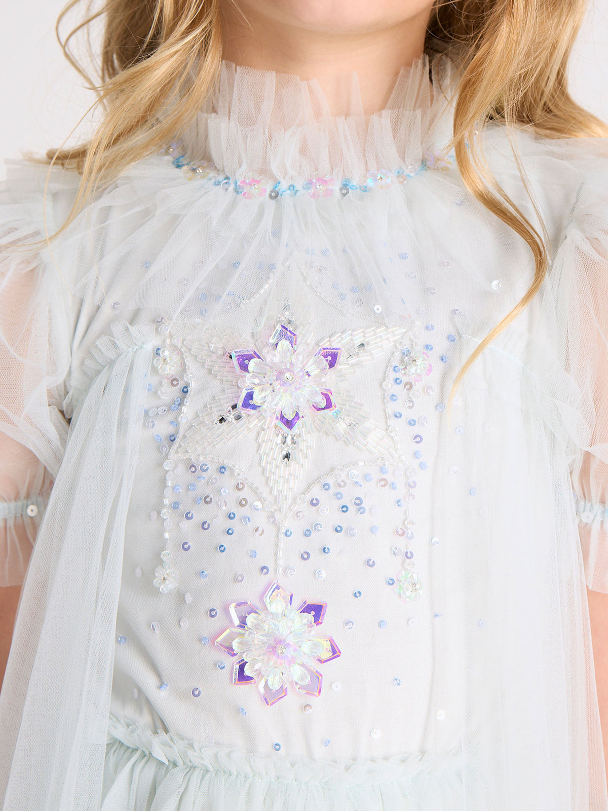 A child wearing the Tutu du Monde Girls’ Snow Queen Tutu Dress, featuring a light blue hue, ruffled collar, iridescent beaded star and flower designs, and sheer tulle skirt details—perfect for an enchanting ice queen costume.