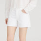 A person in a white long-sleeve blouse with lace cuffs and Frame’s The Long Hang Short, featuring a 4.5” inseam, stands with one hand in their pocket against a plain background for a relaxed look.