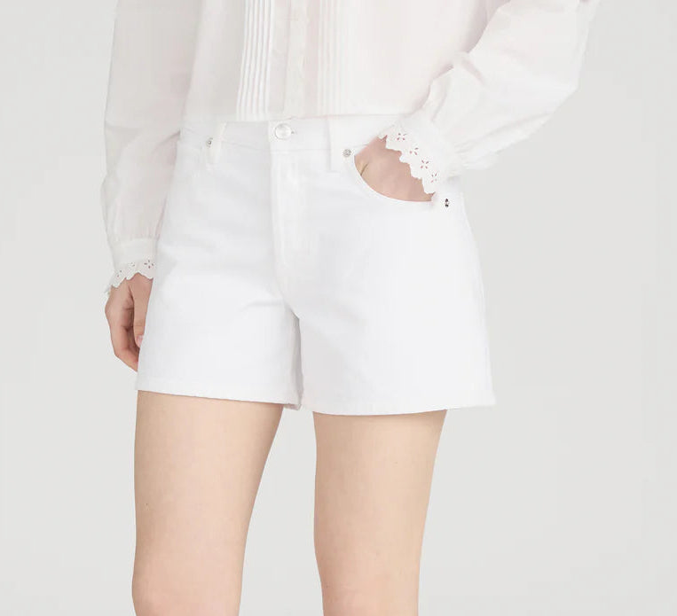 A person in a white long-sleeve blouse with lace cuffs and Frame’s The Long Hang Short, featuring a 4.5” inseam, stands with one hand in their pocket against a plain background for a relaxed look.
