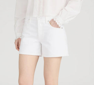 A person in a white long-sleeve blouse with lace cuffs and Frame’s The Long Hang Short, featuring a 4.5” inseam, stands with one hand in their pocket against a plain background for a relaxed look.