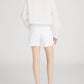 A person stands facing away in a relaxed style, wearing a long-sleeve white blouse, Frame The Long Hang Short with 4½" inseam, and black open-toe heeled sandals against a plain light background.