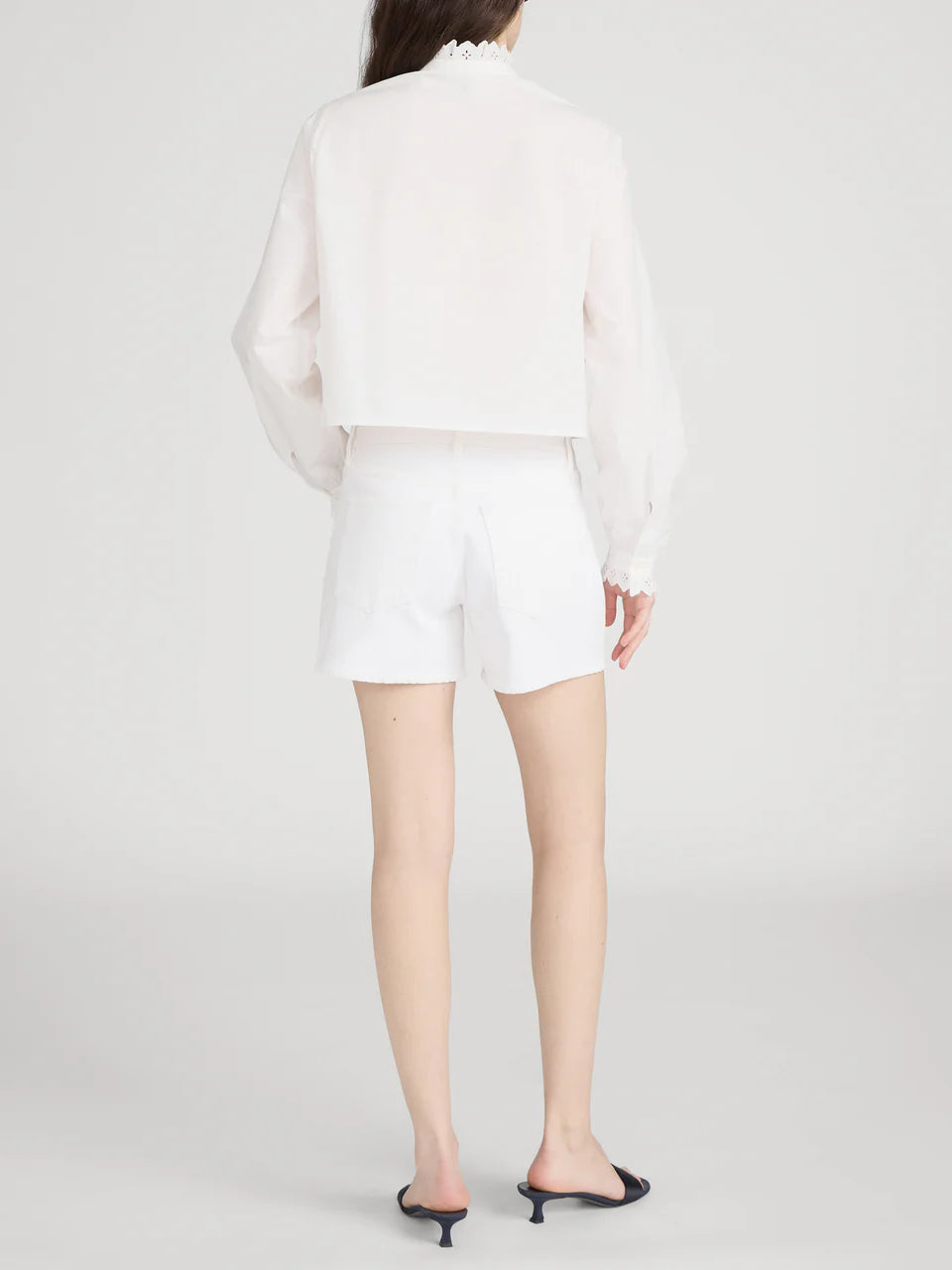 A person stands facing away in a relaxed style, wearing a long-sleeve white blouse, Frame The Long Hang Short with 4½" inseam, and black open-toe heeled sandals against a plain light background.