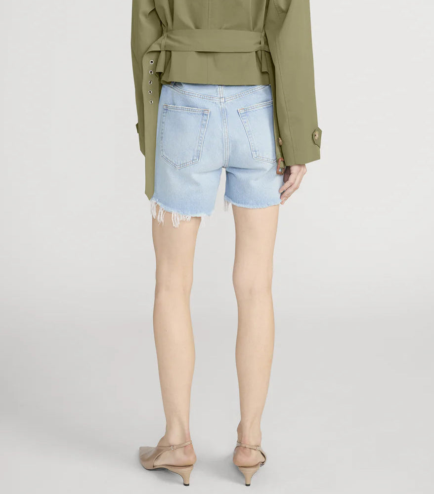 Person wearing a green jacket and Frame’s The Everyday Short—pure cotton, high-rise, 4" inseam with frayed hems—paired with beige low-heeled shoes, standing against a plain background, viewed from behind.
