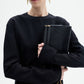 A woman in the CO Keyhole Dress, a black long-sleeved piece, holds a textured black clutch with a gold zipper and chain against a plain white background.