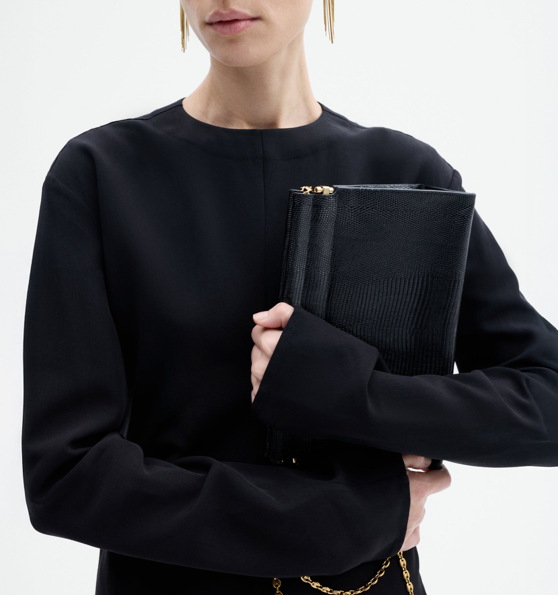A woman in the CO Keyhole Dress, a black long-sleeved piece, holds a textured black clutch with a gold zipper and chain against a plain white background.