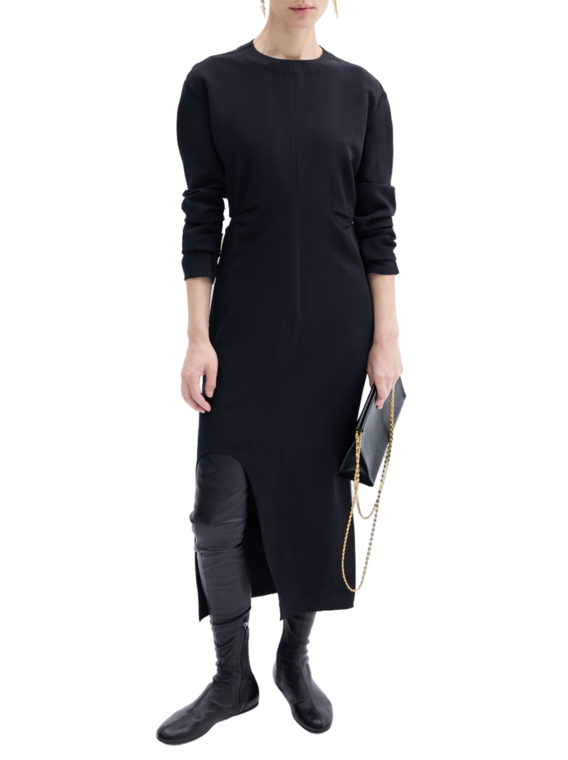 A person wearing CO's minimalist black Keyhole Dress with long sleeves, paired with black boots and holding a black clutch with a gold chain, stands against a plain white background.