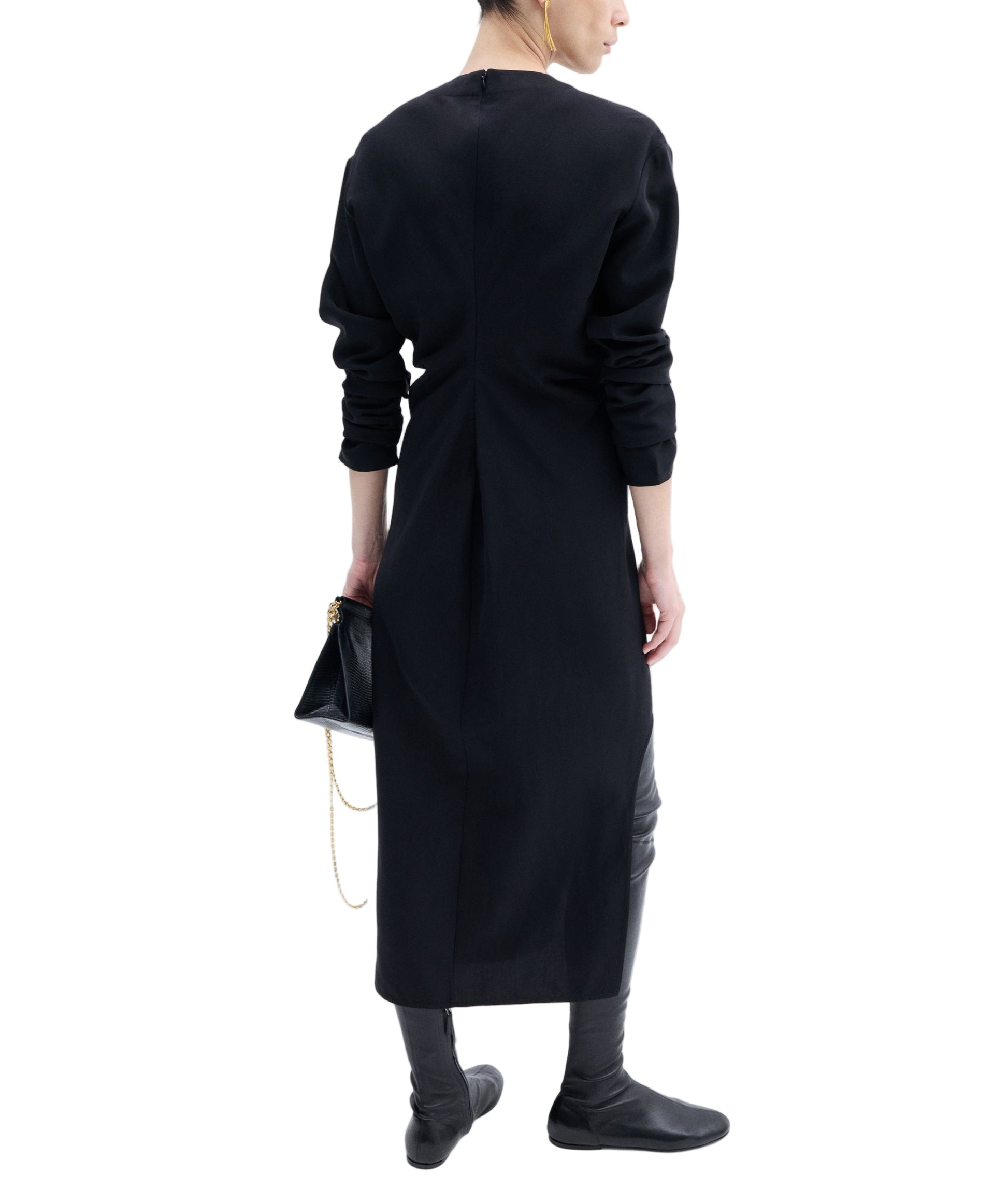 A person stands with their back to the camera, wearing CO's black Keyhole Dress crafted from a viscose wool blend, paired with black shoes and carrying a small black handbag with a gold chain.