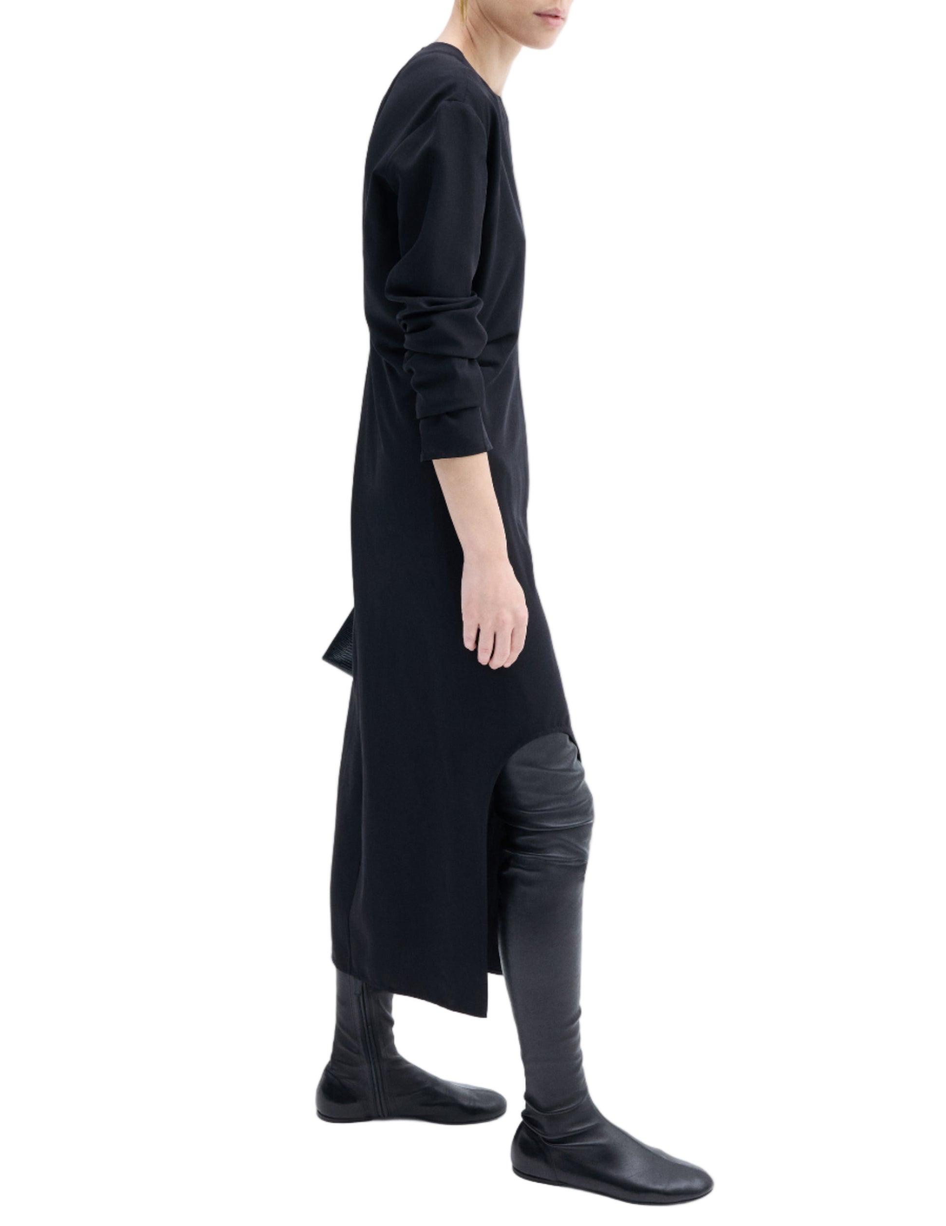A person in a CO Keyhole Dress, minimalist black with long sleeves, paired with black over-the-knee boots, shown in profile against a white background.