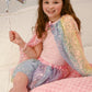 A young girl in a pink top and Great Pretenders Ombre Sequins Skirt with matching cape sits on a pink blanket, smiling as she holds a silver star balloon.