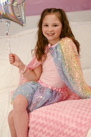 A young girl in a pink top and Great Pretenders Ombre Sequins Skirt with matching cape sits on a pink blanket, smiling as she holds a silver star balloon.