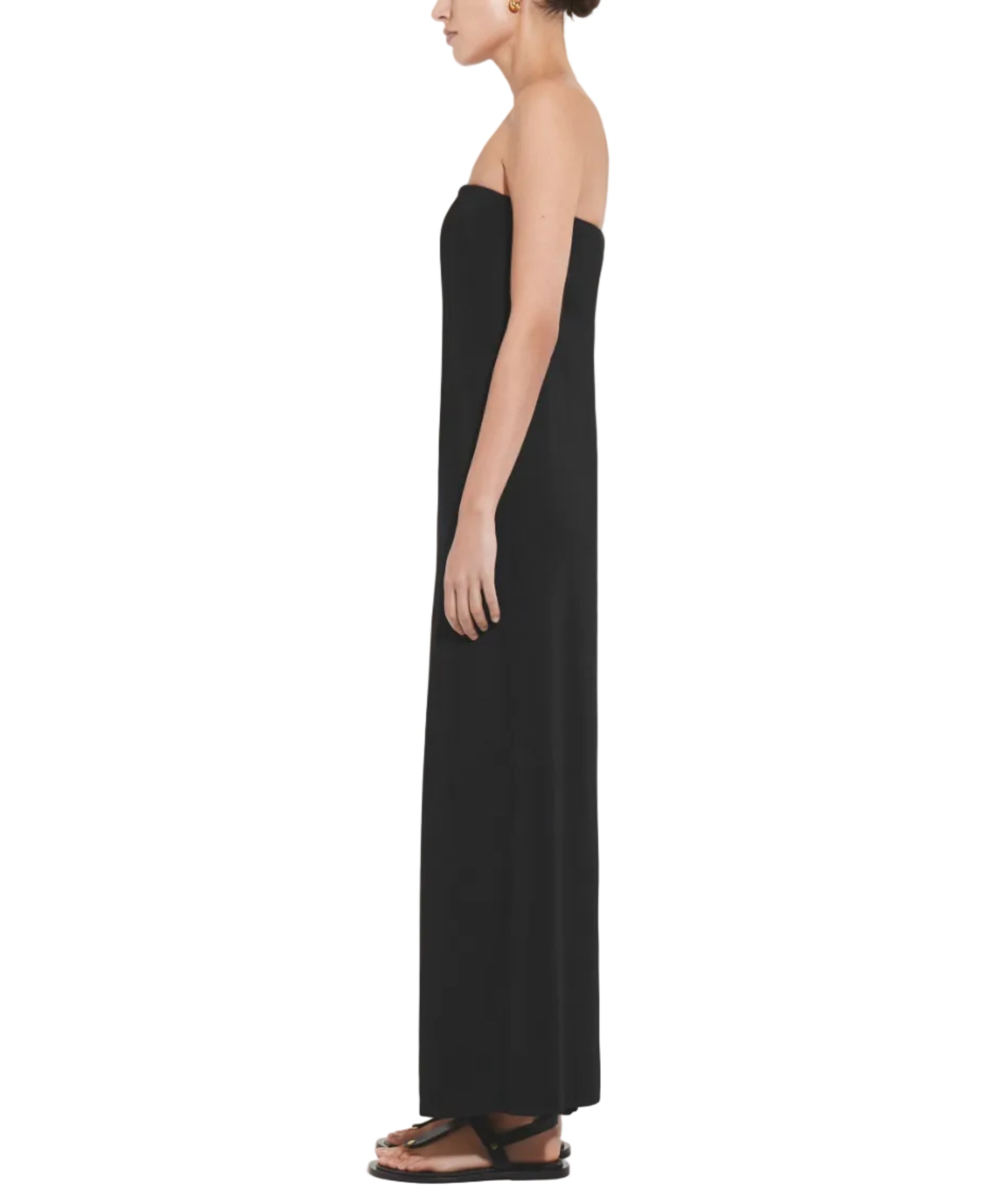 A woman stands in profile wearing the CO Strapless Dress, a full-length black column style paired with black flat sandals—an elegant formal look against a white background.