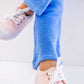 A person wearing light pink sneakers with white laces and The Beaufort Bonnet Company’s Girls’ Terrycloth Patty Pants in light blue, made from soft cotton terrycloth, sits on a white surface with legs crossed at the ankles.