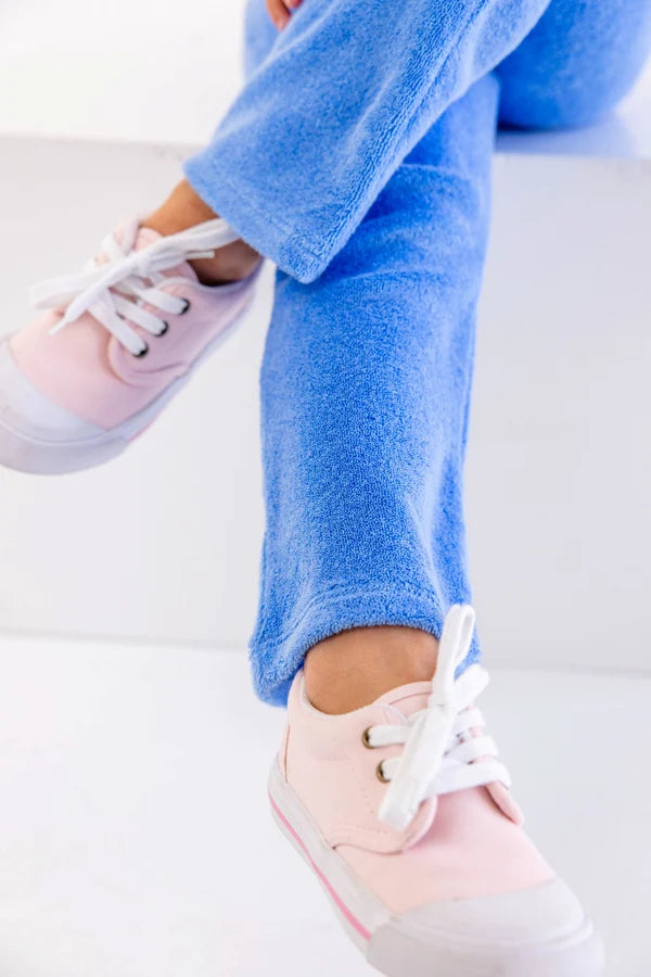 A person wearing light pink sneakers with white laces and The Beaufort Bonnet Company’s Girls’ Terrycloth Patty Pants in light blue, made from soft cotton terrycloth, sits on a white surface with legs crossed at the ankles.