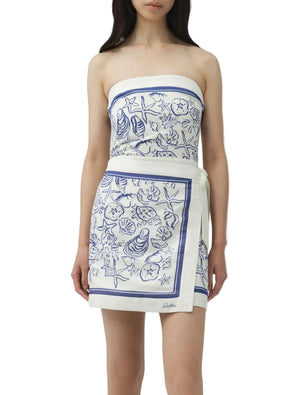 A woman wears the Simkhai Darbie Linen Mini Dress, a white wrap mini dress with blue seashell prints and a matching waist belt.