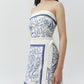 A woman wears the Simkhai Darbie Linen Mini Dress, a bustier mini dress with blue sea-themed seashell motifs, wrap silhouette, and side tie, as she stands against a plain background.