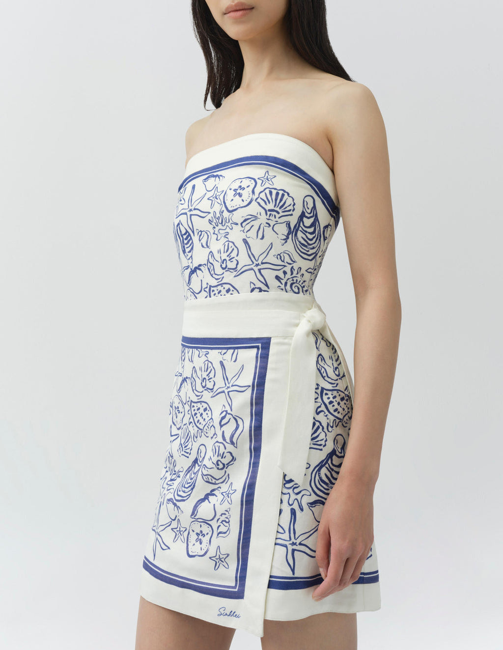 A woman wears the Simkhai Darbie Linen Mini Dress, a bustier mini dress with blue sea-themed seashell motifs, wrap silhouette, and side tie, as she stands against a plain background.